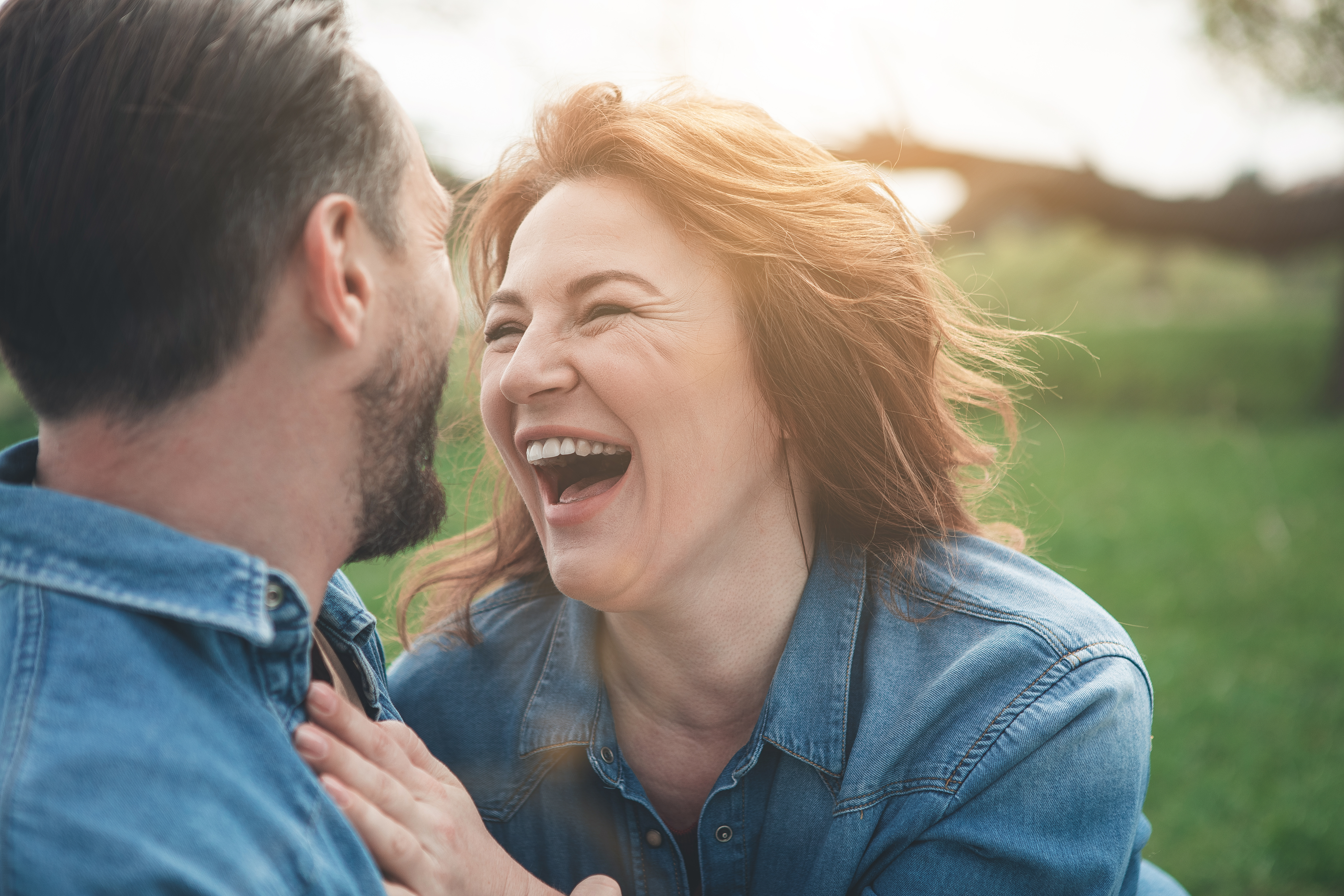 Cherishing Joyful Moments as a Carent – A middle-aged woman and man in denim jackets share a joyful moment outdoors, laughing together in a sunlit park. A theme of finding happiness and connection despite the challenges of caring for an elderly parent