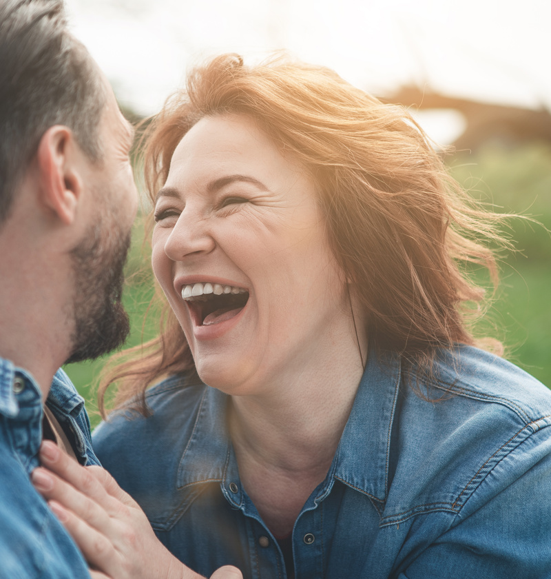 Cherishing Joyful Moments as a Carent – A middle-aged woman and man in denim jackets share a joyful moment outdoors, laughing together in a sunlit park. A theme of finding happiness and connection despite the challenges of caring for an elderly parent