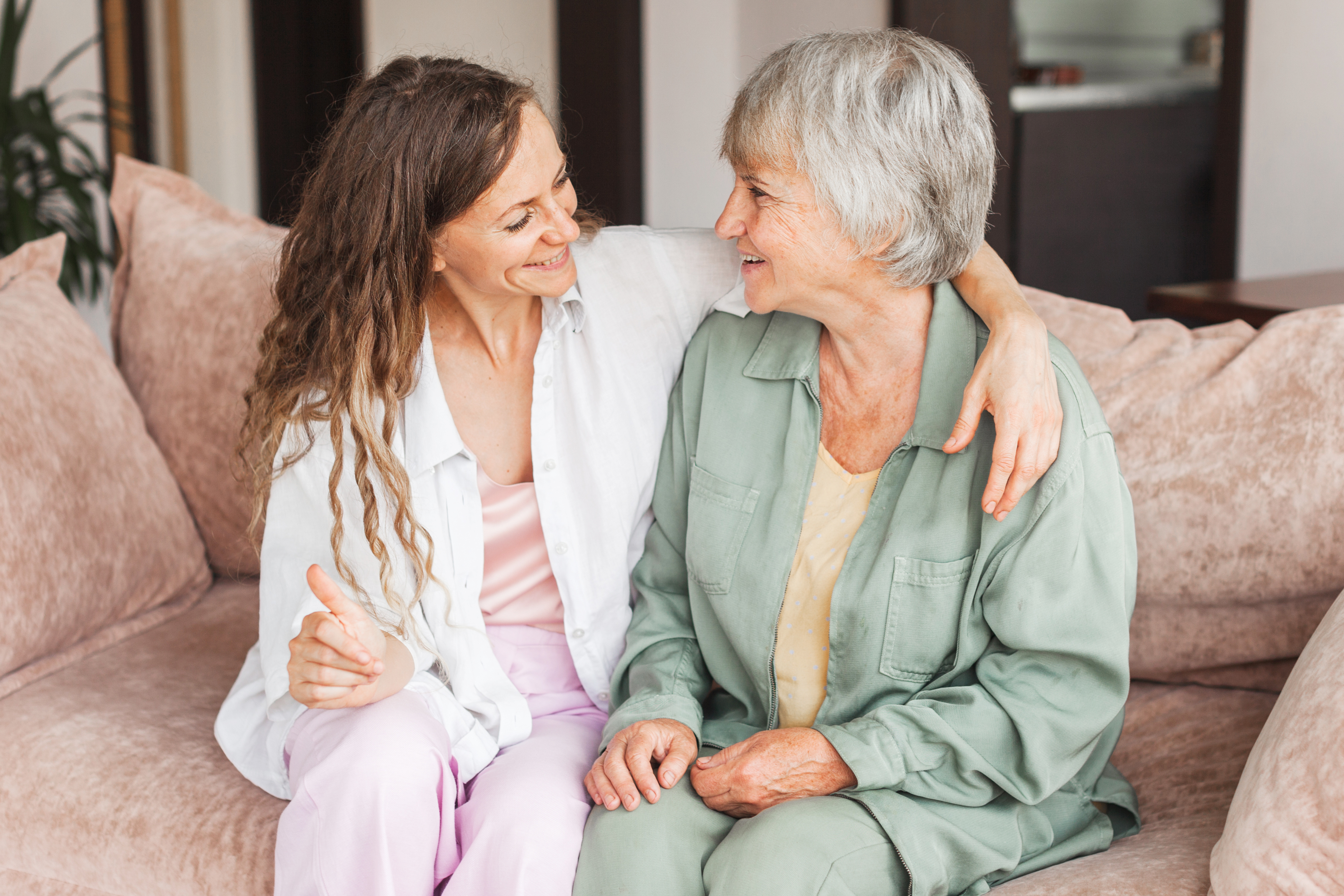 A smiling adult daughter sits close to her elderly mother on a sofa, sharing a warm moment and holding hands. They are looking at each other with affection, showing a strong, supportive relationship.
