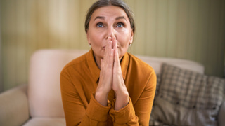 Finding Hope and Strength as a Carent When Feeling Frustrated – A woman in her 50s or 60s wearing a mustard-coloured blouse sits on a sofa, gazing upwards with clasped hands. A moment of resilience, reflection, and finding inner strength in the caregiving journey.