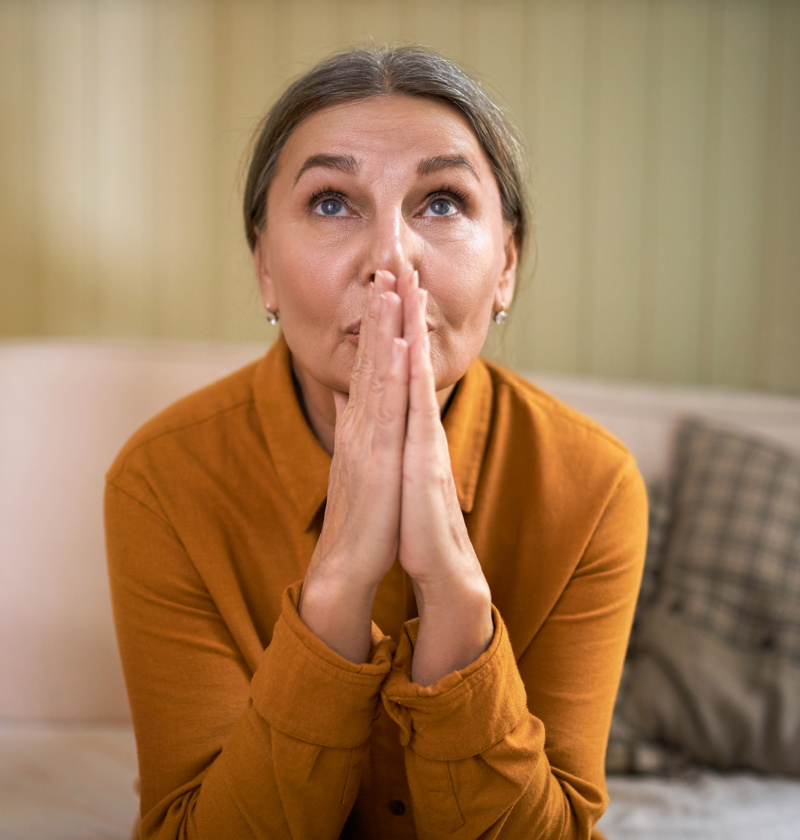 Finding Hope and Strength as a Carent When Feeling Frustrated – A woman in her 50s or 60s wearing a mustard-coloured blouse sits on a sofa, gazing upwards with clasped hands. A moment of resilience, reflection, and finding inner strength in the caregiving journey.