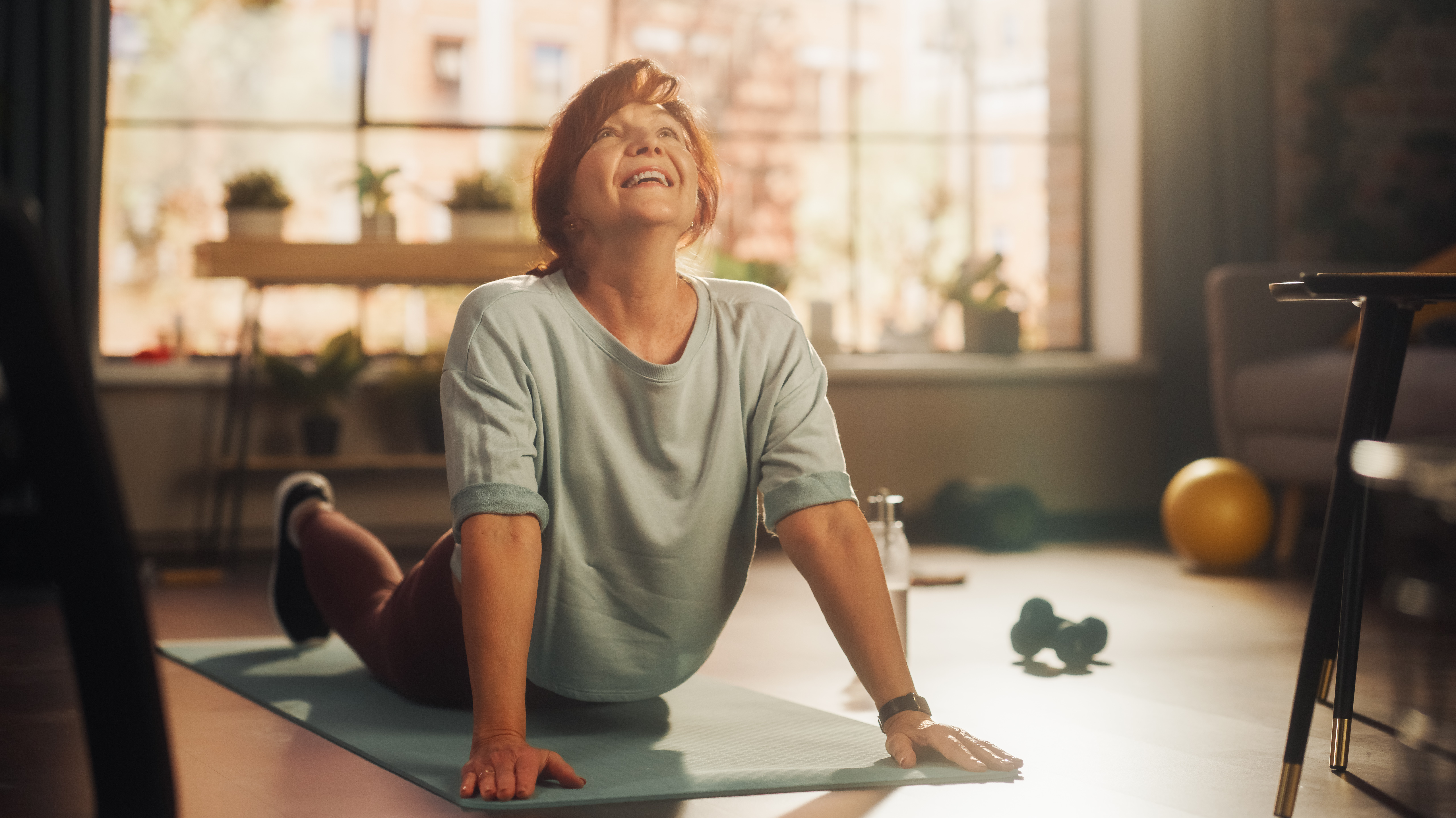 Self-Care and Wellbeing for Carents – A smiling woman in her 50s or 60s does a yoga stretch on a mat in a bright, cosy living room. A representation of self-care, stress relief, and maintaining wellbeing while caring for an elderly parent.