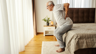 Older man sitting up from bed with visible back discomfort – highlighting the need for bed sensor pads to monitor movement, alert carers to changes, and support fall prevention for elderly parents.