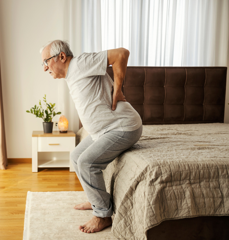 Older man sitting up from bed with visible back discomfort – highlighting the need for bed sensor pads to monitor movement, alert carers to changes, and support fall prevention for elderly parents.
