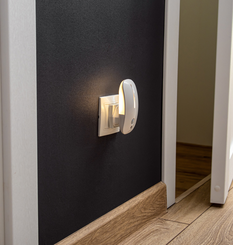 Plug-in motion sensor night light switched on in a hallway – helps elderly parents move safely around the home at night by automatically lighting dark areas without needing to search for switches.