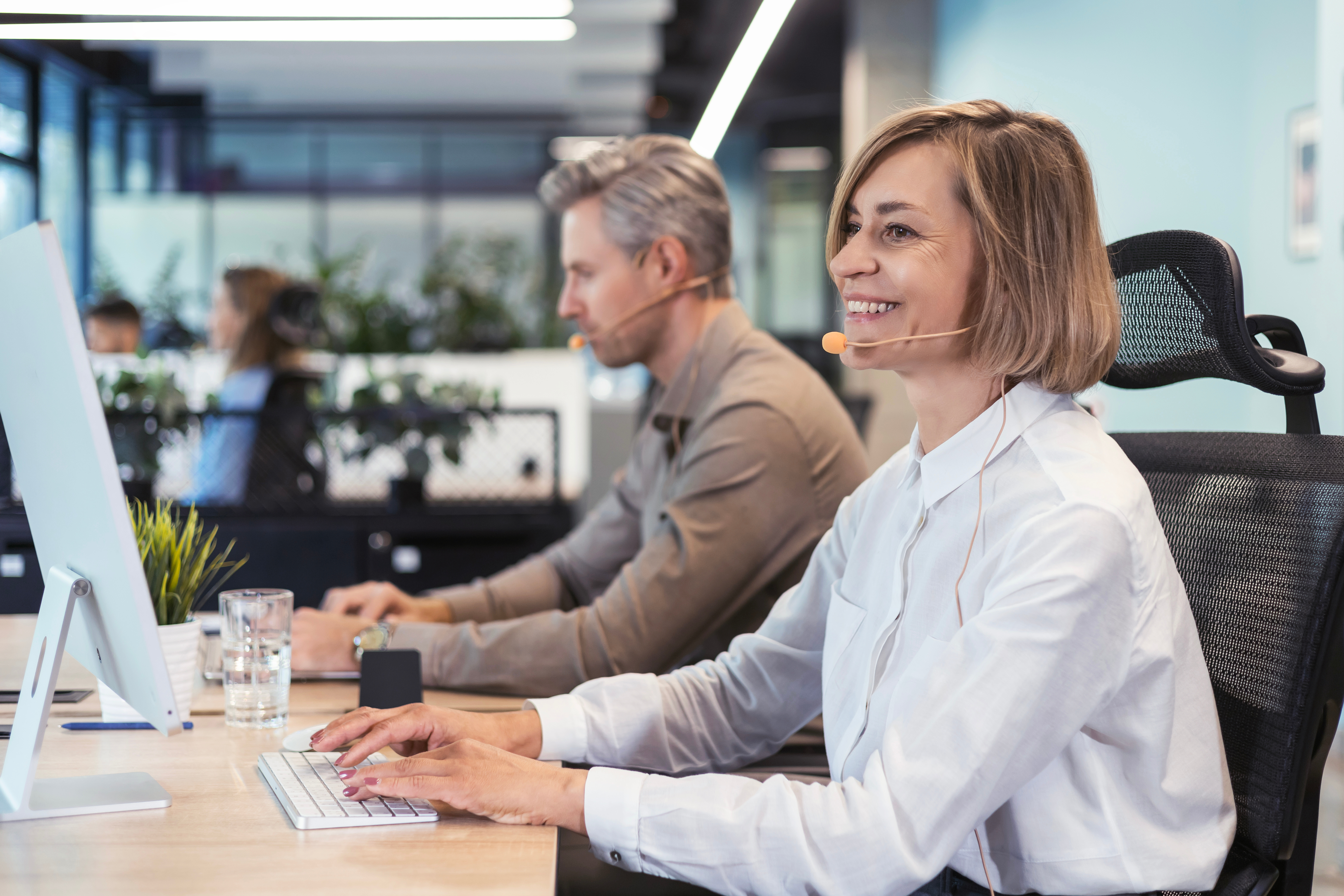 A smiling woman wearing a headset types on a keyboard in a bright office. A representation of working carers balancing employment with caring responsibilities and understanding workplace rights, flexible working, and carer support policies.