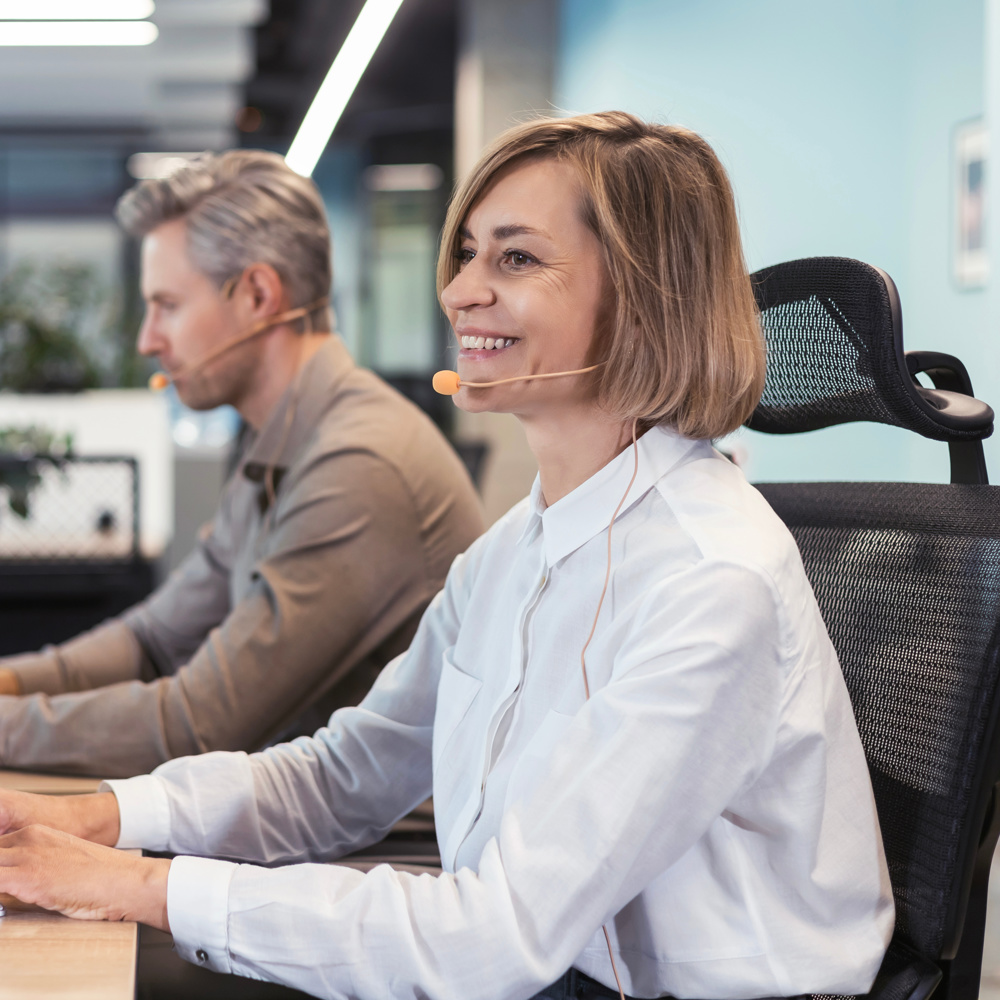 A smiling woman wearing a headset types on a keyboard in a bright office. A representation of working carers balancing employment with caring responsibilities and understanding workplace rights, flexible working, and carer support policies.
