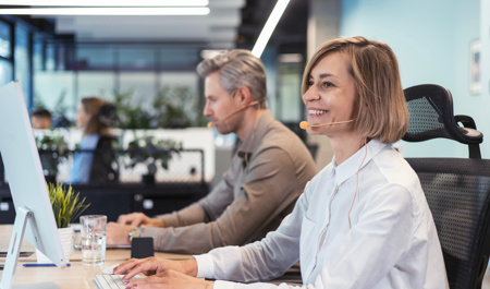 A smiling woman wearing a headset types on a keyboard in a bright office. A representation of working carers balancing employment with caring responsibilities and understanding workplace rights, flexible working, and carer support policies.