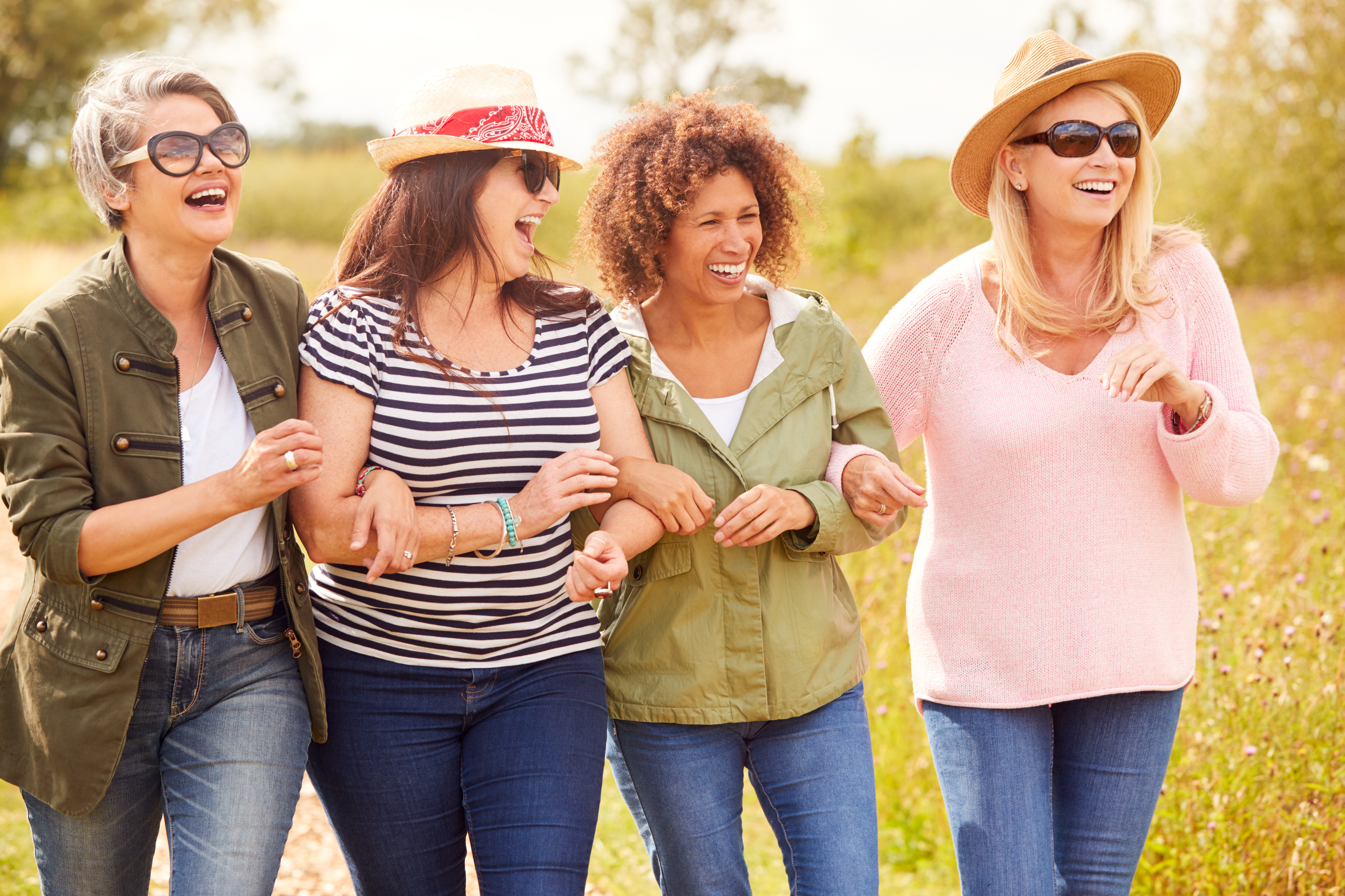  A group of four middle-aged women walking arm in arm through a countryside path, smiling and laughing. A reminder of the importance of friendship and emotional support for those caring for elderly parents.