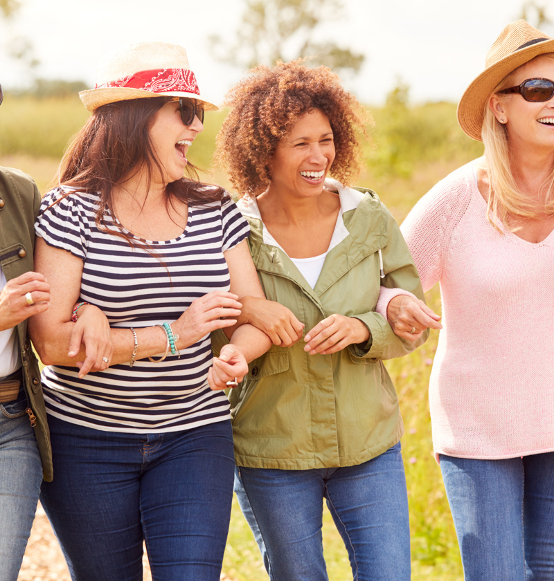  A group of four middle-aged women walking arm in arm through a countryside path, smiling and laughing. A reminder of the importance of friendship and emotional support for those caring for elderly parents.