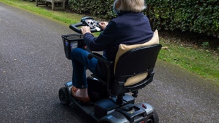 Elderly parent using a mobility scooter outdoors on a paved path – mobility scooters offer elderly parents greater independence and comfort when travelling short distances outside the home.