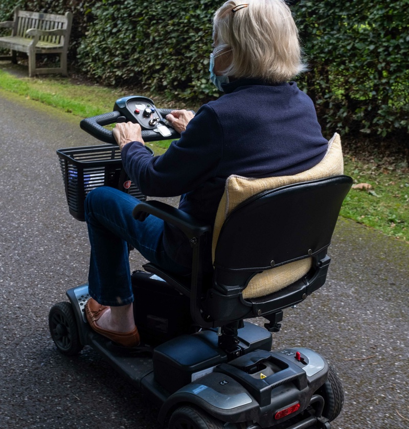 Elderly parent using a mobility scooter outdoors on a paved path – mobility scooters offer elderly parents greater independence and comfort when travelling short distances outside the home.