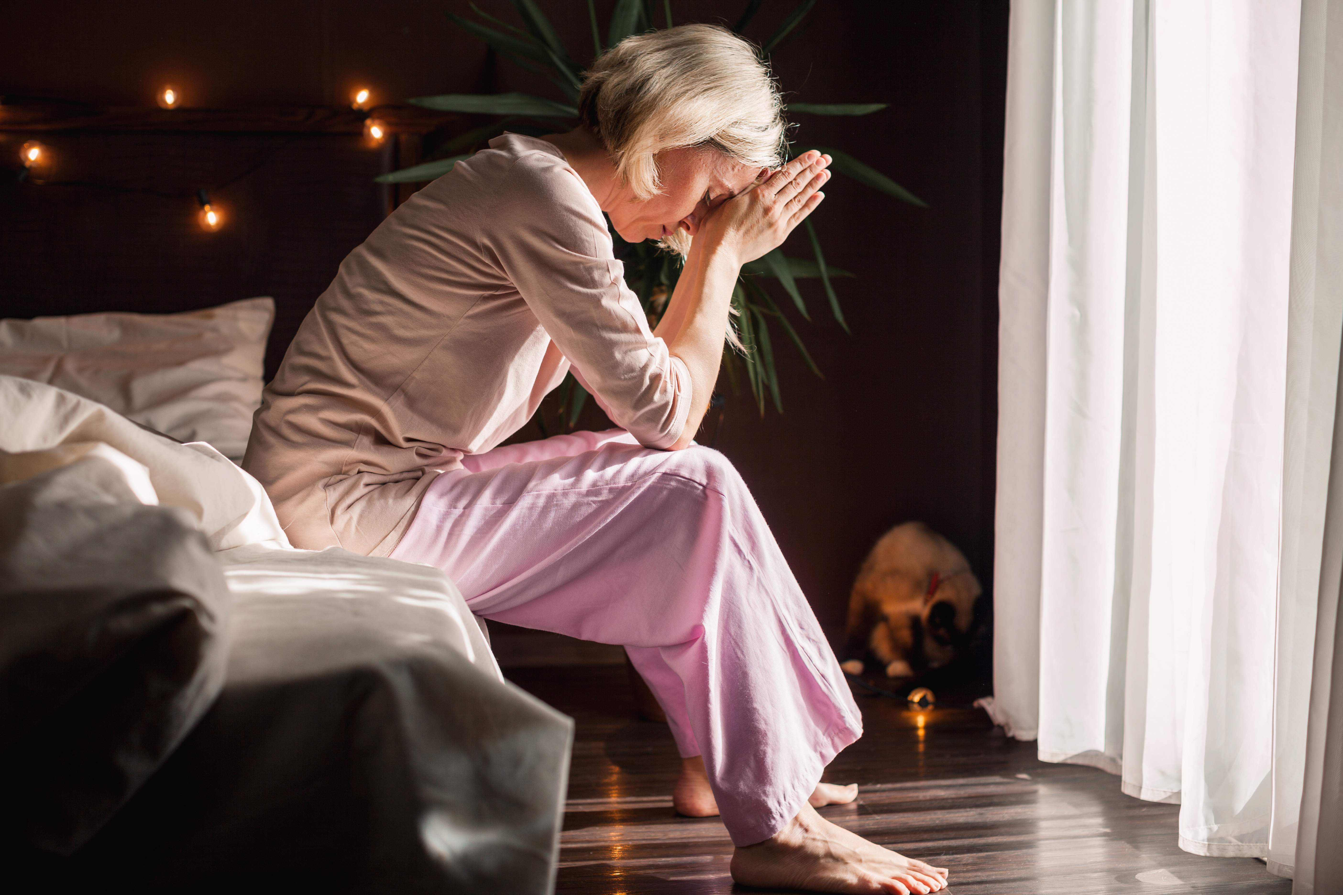 Caring for a Parent While Managing Stress – A middle-aged woman in pink pyjamas sits on the edge of her bed, deep in thought, hands clasped near her face. A portrayal of the emotional toll and mental fatigue that can come with caring for an ageing parent.