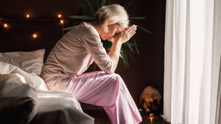 Caring for a Parent While Managing Stress – A middle-aged woman in pink pyjamas sits on the edge of her bed, deep in thought, hands clasped near her face. A portrayal of the emotional toll and mental fatigue that can come with caring for an ageing parent.