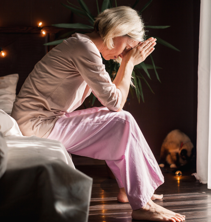 Caring for a Parent While Managing Stress – A middle-aged woman in pink pyjamas sits on the edge of her bed, deep in thought, hands clasped near her face. A portrayal of the emotional toll and mental fatigue that can come with caring for an ageing parent.