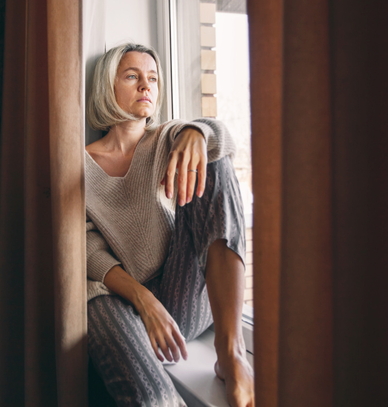 Reflecting on the Challenges of Caring for an eldery parent – A middle-aged woman in a warm sweater and patterned pyjama bottoms gazes out of a window with a pensive expression. A depiction of the isolation and quiet moments of reflection experienced by many caring for elderly parents.