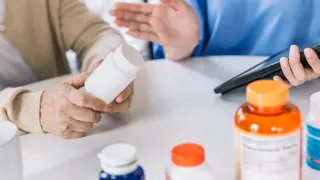 Older person holding a bottle of pills, with many other containers of pills around 