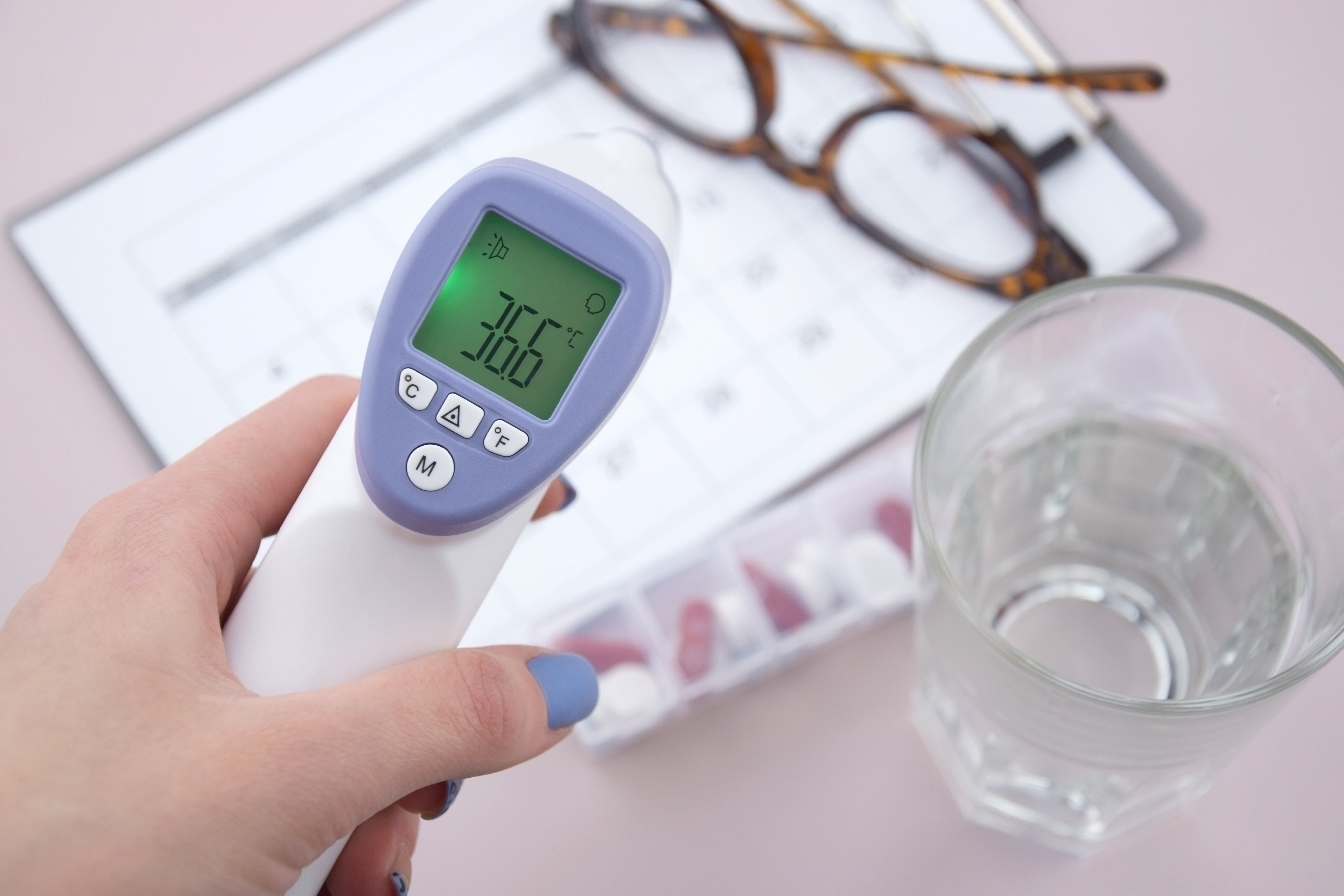 Digital forehead thermometer showing 36.6°C, used as part of daily health checks for elderly parents. Seen with a pill organiser, water glass, and care schedule — ideal for carers monitoring temperature and medication at home.