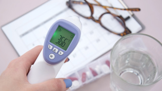 Digital forehead thermometer showing 36.6°C, used as part of daily health checks for elderly parents. Seen with a pill organiser, water glass, and care schedule — ideal for carers monitoring temperature and medication at home.