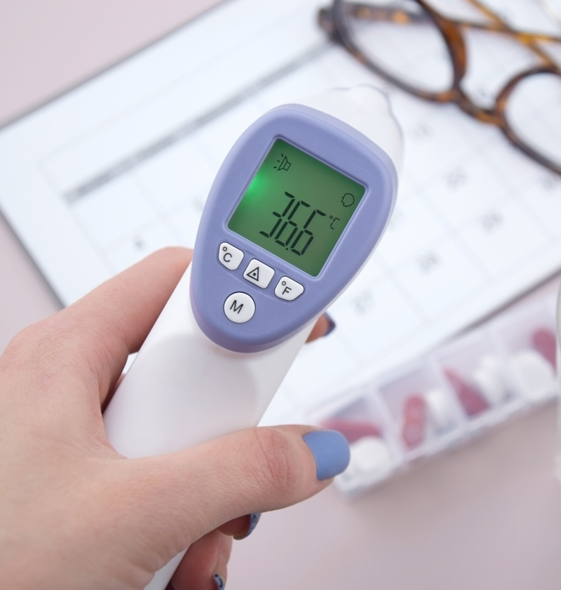 Digital forehead thermometer showing 36.6°C, used as part of daily health checks for elderly parents. Seen with a pill organiser, water glass, and care schedule — ideal for carers monitoring temperature and medication at home.