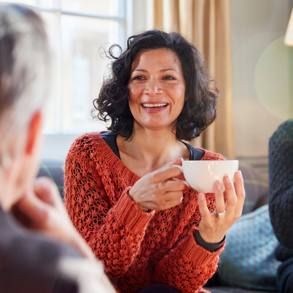 Support Networks: Carents Connecting Over Coffee – A cheerful woman in a burnt-orange knitted sweater holds a cup of coffee, engaging in a warm conversation with friends. A representation of emotional support, carer connections, and the importance of social interaction while navigating caregiving responsibilities.