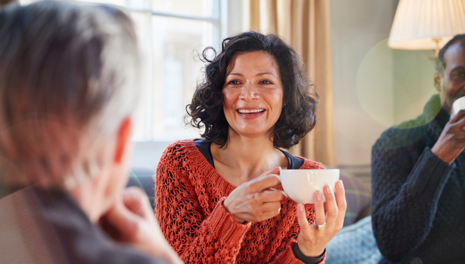 Support Networks: Carents Connecting Over Coffee – A cheerful woman in a burnt-orange knitted sweater holds a cup of coffee, engaging in a warm conversation with friends. A representation of emotional support, carer connections, and the importance of social interaction while navigating caregiving responsibilities.