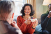Support Networks: Carents Connecting Over Coffee – A cheerful woman in a burnt-orange knitted sweater holds a cup of coffee, engaging in a warm conversation with friends. A representation of emotional support, carer connections, and the importance of social interaction while navigating caregiving responsibilities.