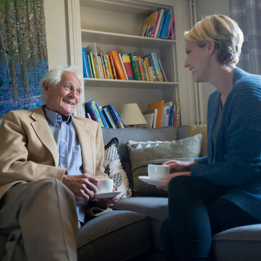 Image of an elderly parent at home drinking a cup of tea, happily sitting on a couch 