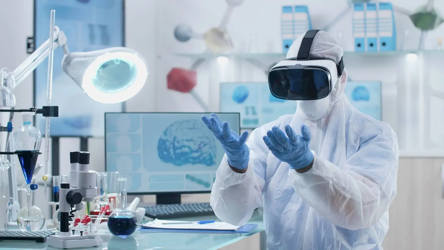 Scientist wearing a VR machine looking at his hands - behind him we can see a picture of a brain scan 