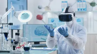 Scientist wearing a VR machine looking at his hands - behind him we can see a picture of a brain scan 