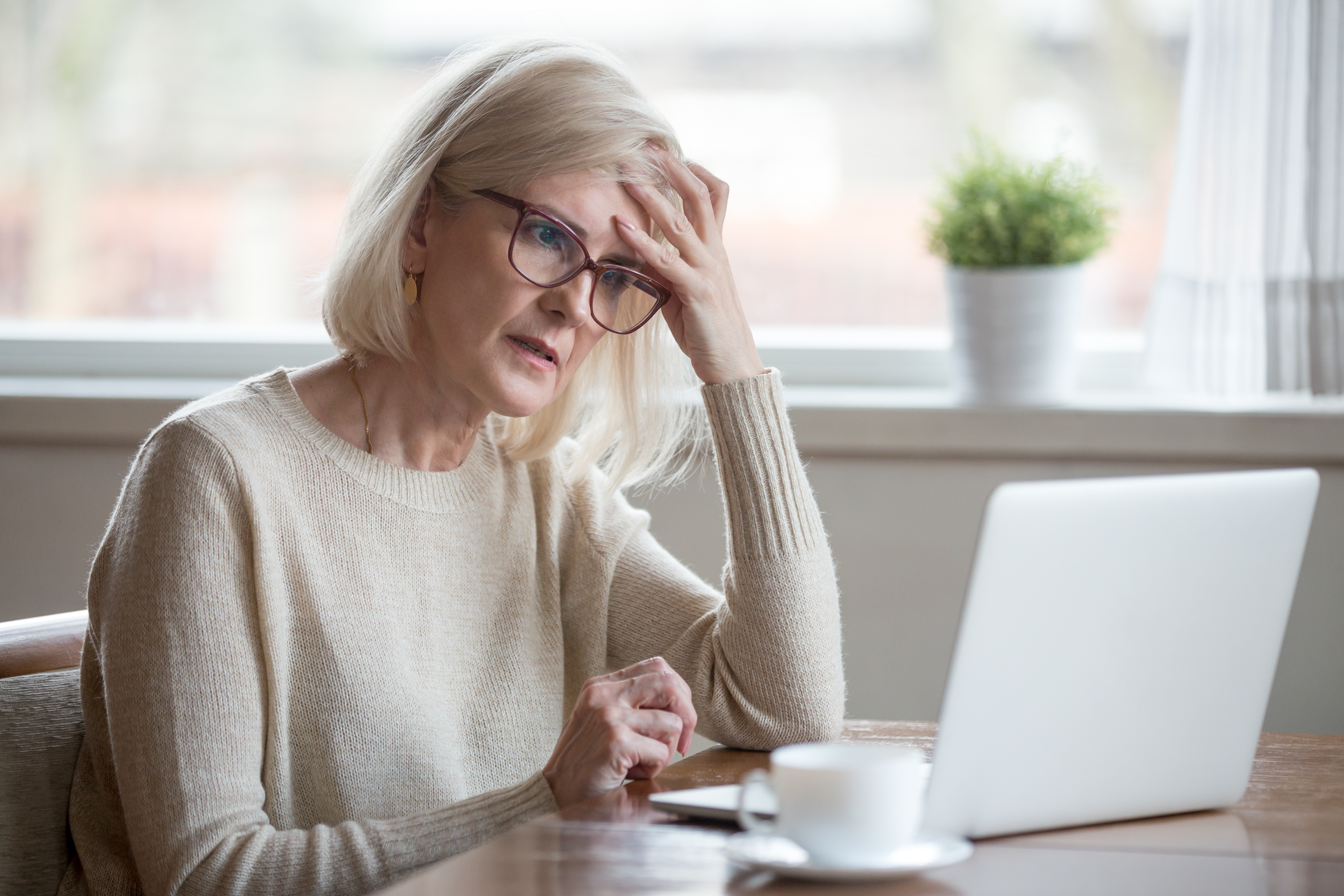 Managing Feelings of Loss While Caring for an Elderly Parent – An older woman in glasses and a beige sweater sits at a table with a laptop, hand on forehead, looking concerned. A depiction of the loss she feels stress that comes with caring for a parent.