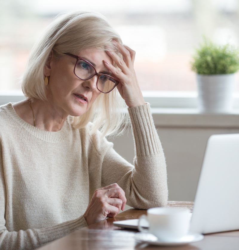 Managing Feelings of Loss While Caring for an Elderly Parent – An older woman in glasses and a beige sweater sits at a table with a laptop, hand on forehead, looking concerned. A depiction of the loss she feels stress that comes with caring for a parent.