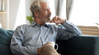 Finding Moments to reflect on your Emotions as a Carent – A silver-haired man sits on a plush sofa, holding a coffee cup while staring thoughtfully into the distance. A representation of taking time for oneself amidst the responsibilities of supporting an ageing parent.