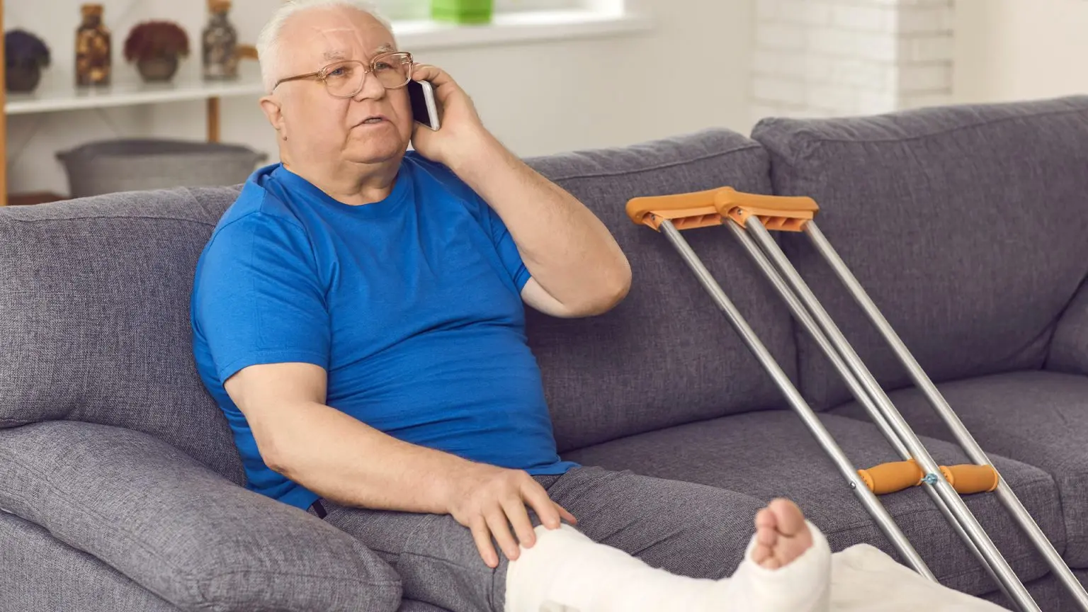 Older gentlemen sitting on the couch with his broken foot up, with a pair of crutches next to him 