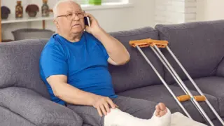 Older gentlemen sitting on the couch with his broken foot up, with a pair of crutches next to him 