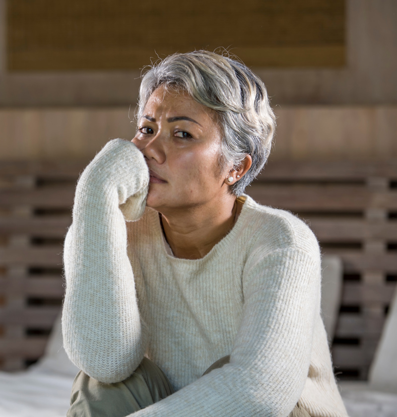 Navigating Stress and Change in Caregiving – A middle-aged woman with salt-and-pepper hair in a cream sweater sits on her bed, resting her hand on her mouth in concern. A scene highlighting the emotional complexities of caring for a parent and coping with change.