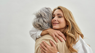 A woman embraces an elderly parent, both smiling gently as they share a moment of affection. The image conveys warmth, care, and emotional connection. The background is soft and neutral, focusing on the bond between them.