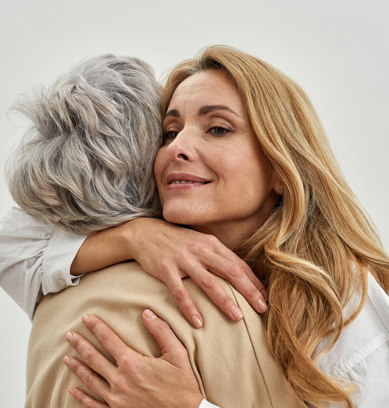 A woman embraces an elderly parent, both smiling gently as they share a moment of affection. The image conveys warmth, care, and emotional connection. The background is soft and neutral, focusing on the bond between them.