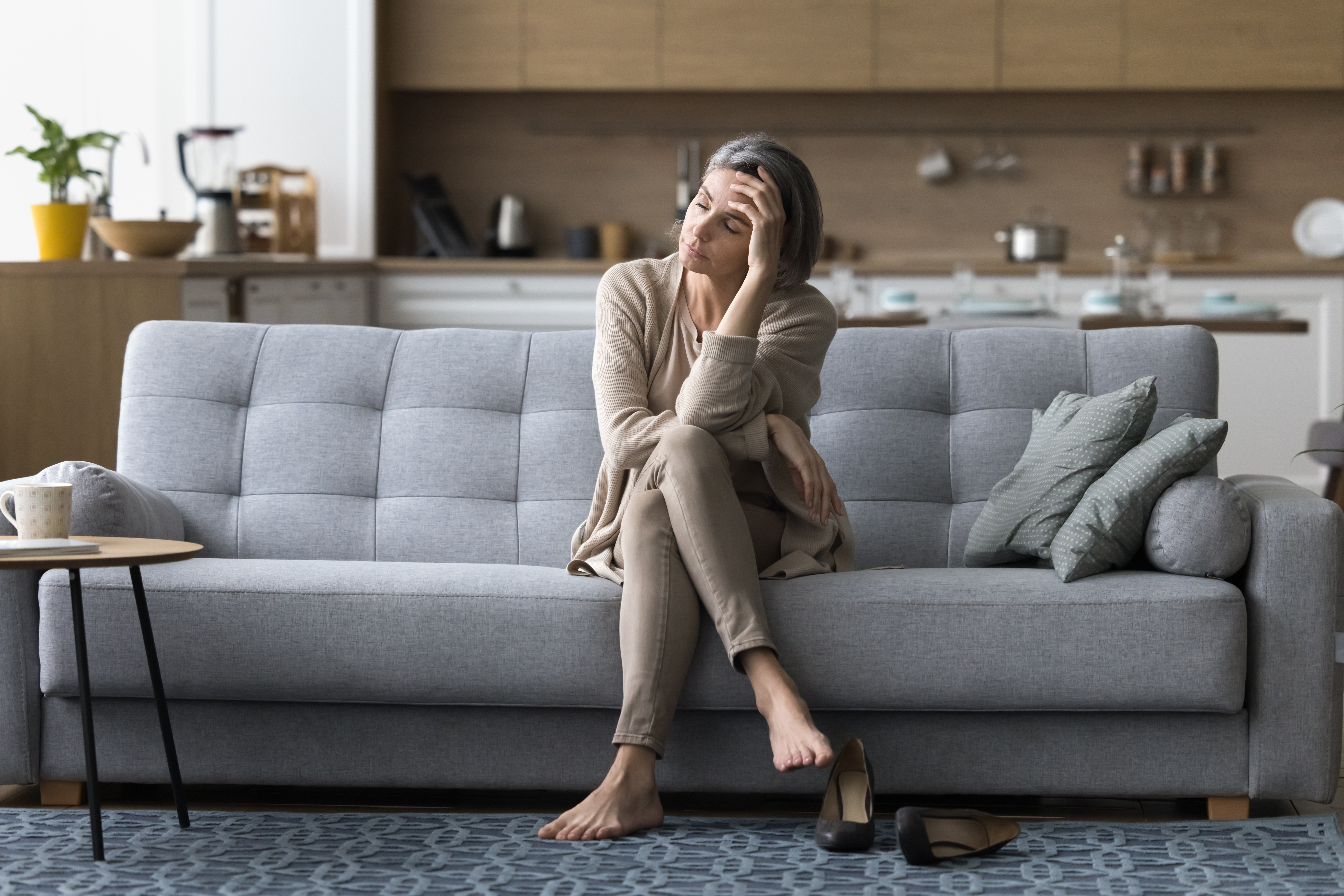 Middle-aged woman looking exhausted and stressed while sitting on a grey sofa at home, symbolising burnout from caring for an elderly relative. Barefoot with shoes off, she appears overwhelmed, highlighting the emotional and physical toll of caring for elderly parents