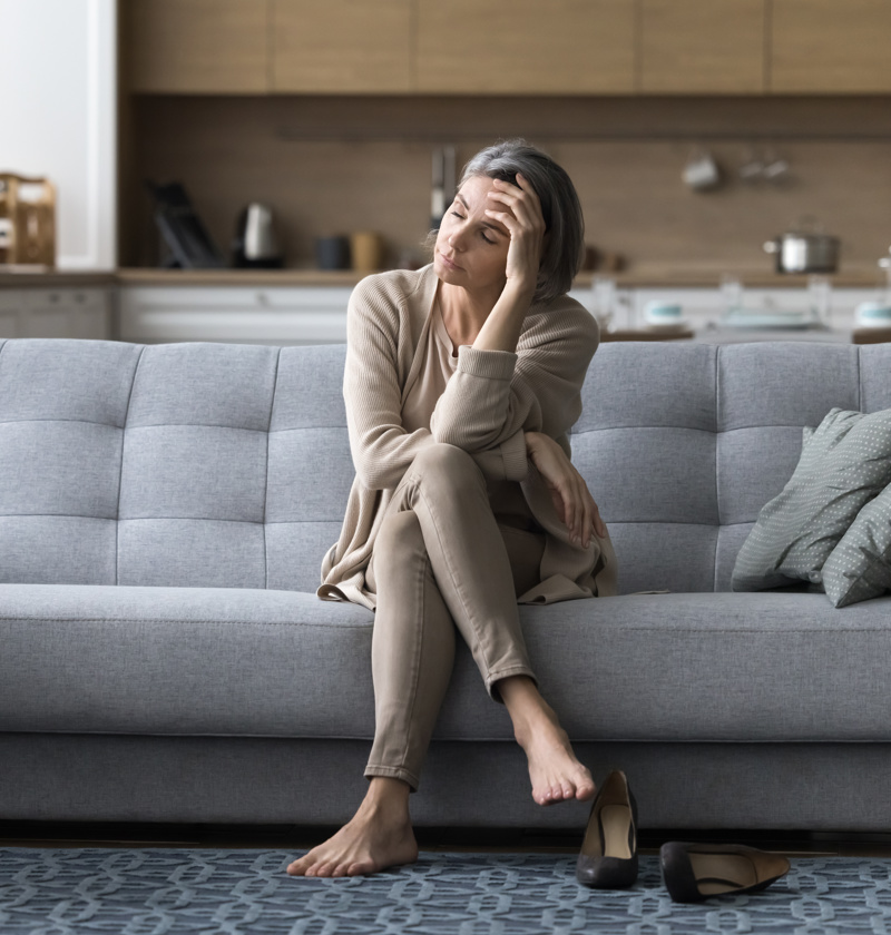 Middle-aged woman looking exhausted and stressed while sitting on a grey sofa at home, symbolising burnout from caring for an elderly relative. Barefoot with shoes off, she appears overwhelmed, highlighting the emotional and physical toll of caring for elderly parents
